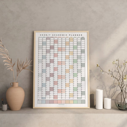 A framed 2026/2027 academic planner poster displayed on a shelf in a styled interior setting with vases, candles, and dried branches against a textured gray wall.