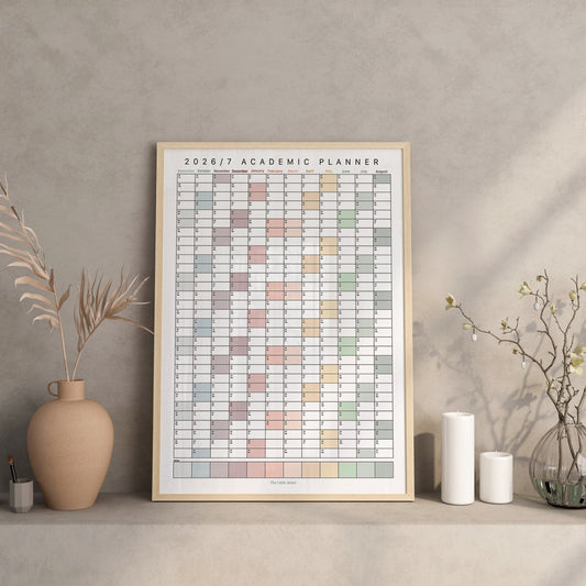 A framed 2026/2027 academic planner poster displayed on a shelf in a styled interior setting with vases, candles, and dried branches against a textured gray wall.
