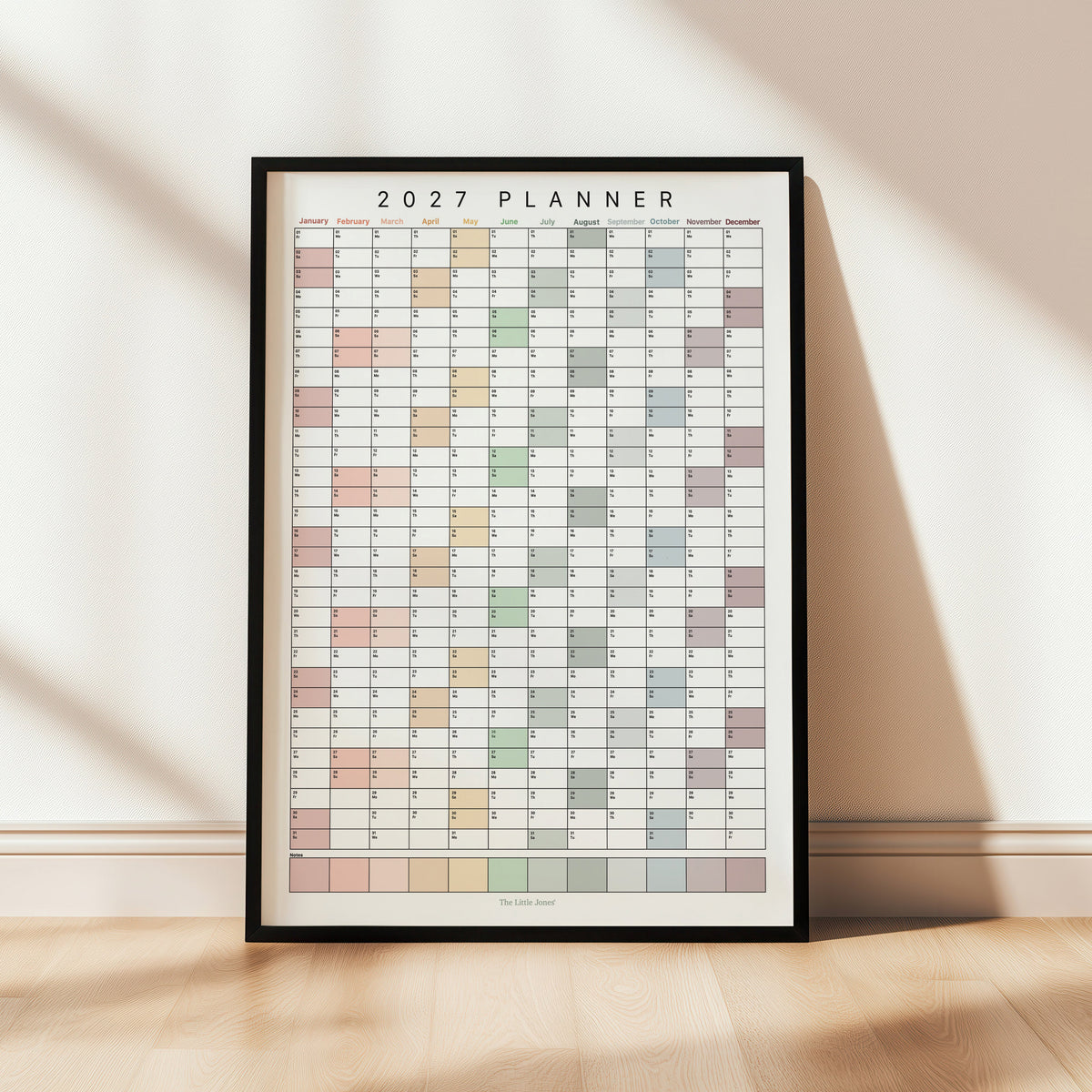 A framed 2027 wall planner poster leaning against a wall in a thin black frame.