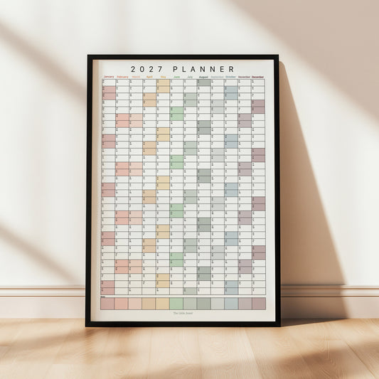 A framed 2027 wall planner poster leaning against a wall in a thin black frame.