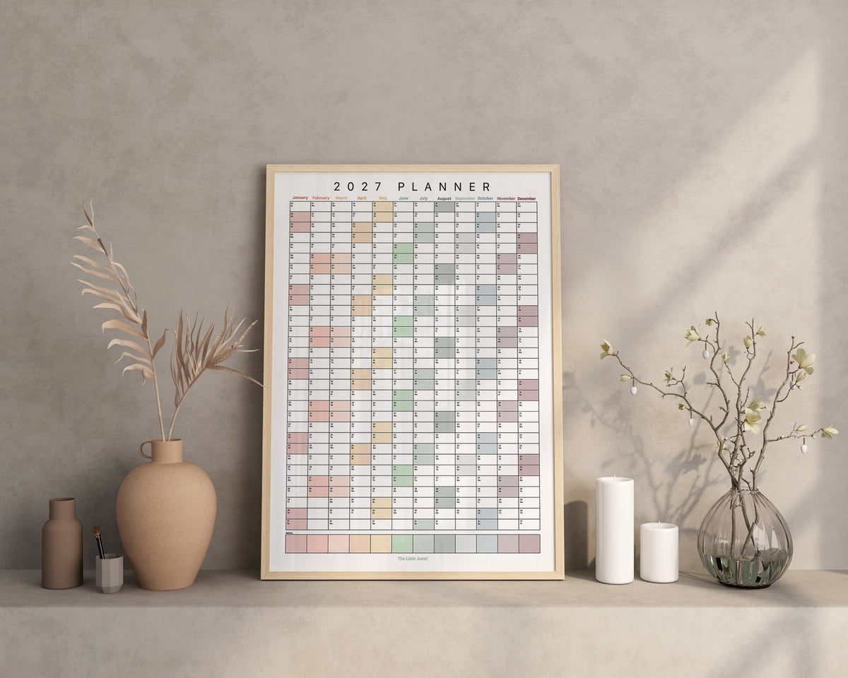 A framed 2027 family wall planner displayed on a shelf against a textured wall, styled with ceramic vases, candles, and dried branches