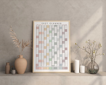 A framed 2027 family wall planner displayed on a shelf against a textured wall, styled with ceramic vases, candles, and dried branches