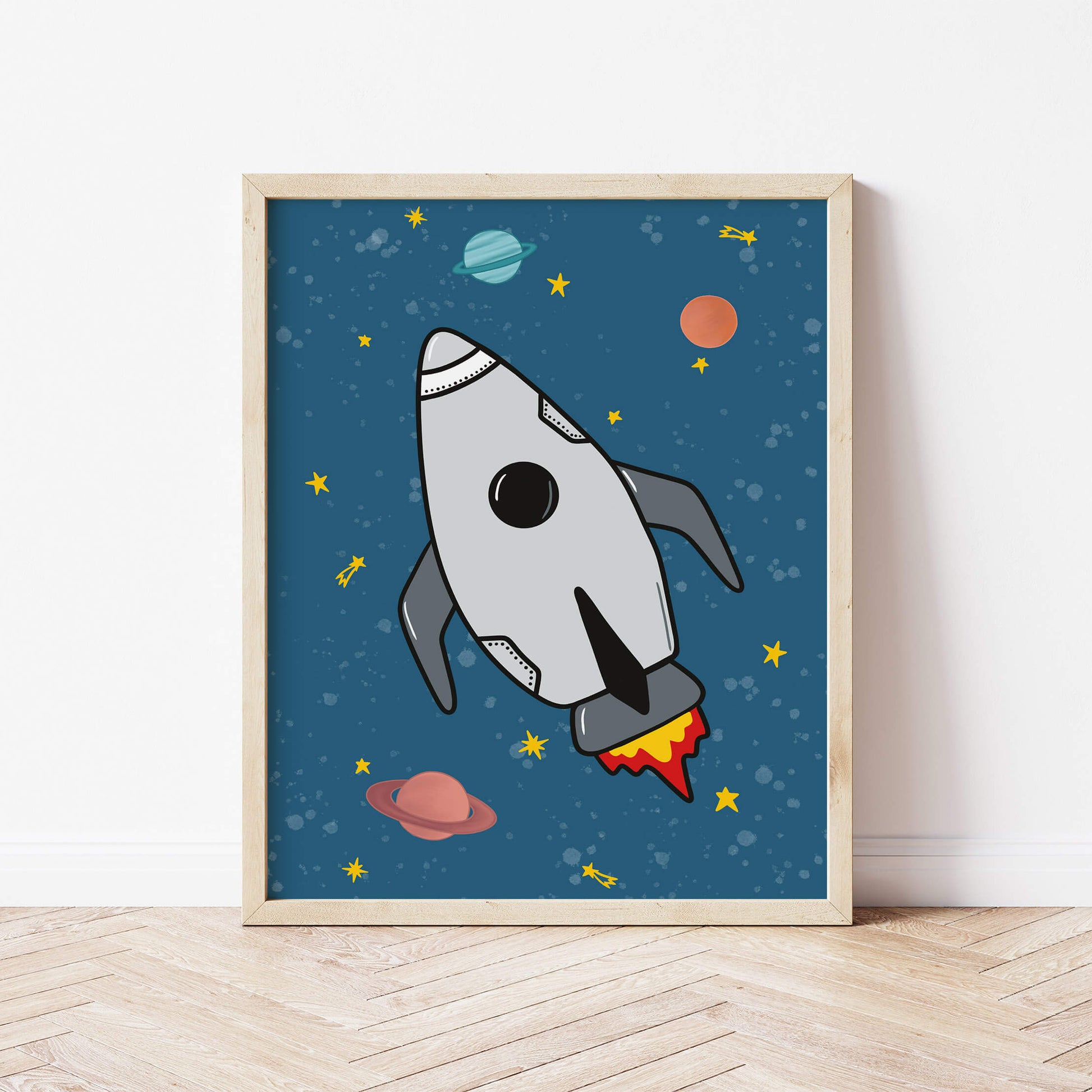 Framed artwork of a rocket ship on a blue background with planets and stars, displayed on a wooden floor.
