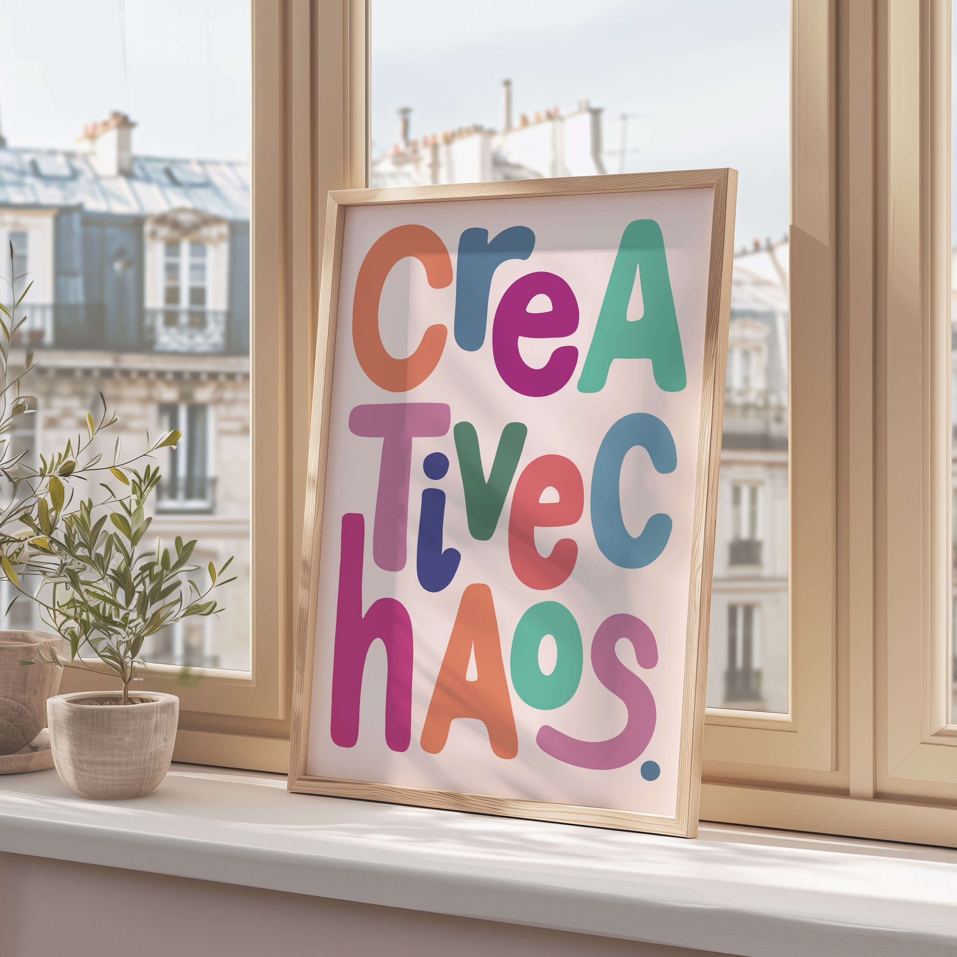 Framed colorful poster with 'Creative Chaos' on a windowsill with cityscape view