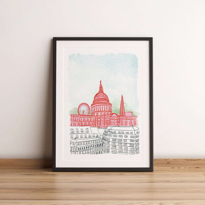 Framed artwork of a cityscape with a prominent building on a wooden floor against a white wall.