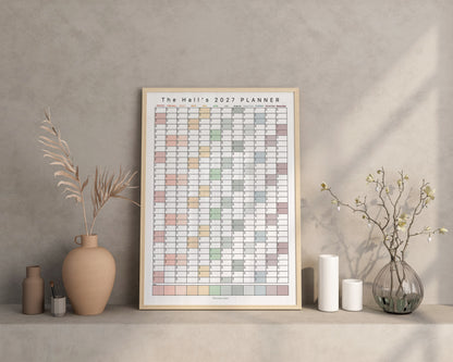 A personalised 2027 wall planner poster displayed on a shelf against a textured gray wall, styled with ceramic vases, candles, and dried branches.